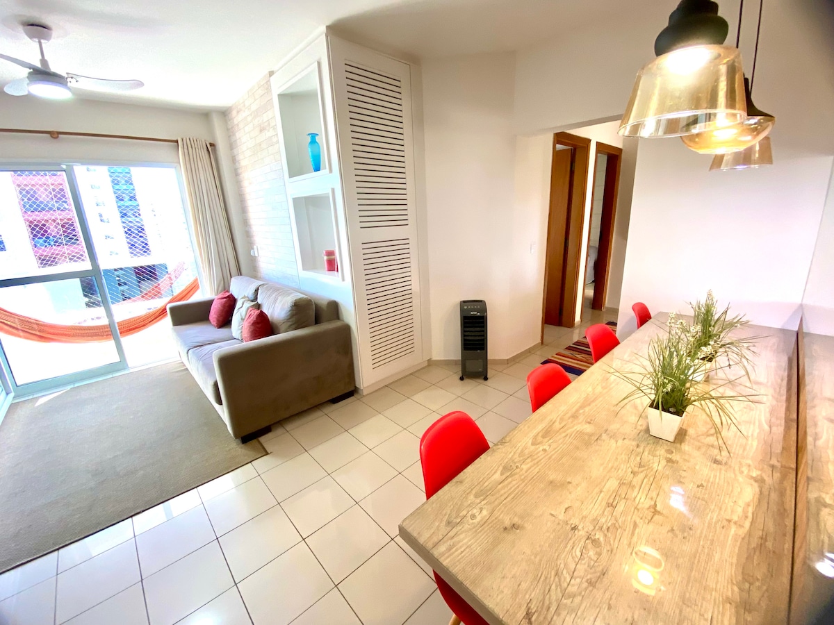 A light-filled living and dining area features a wooden table with red chairs. A cozy sofa is positioned nearby, creating a welcoming space. A sliding glass door opens to a balcony, with a hammock visible outside, providing a connection to the outdoors.