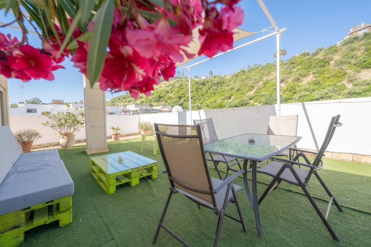 An outdoor terrace features a turf floor and a dining table surrounded by six chairs. A comfortable seating area is arranged with a sofa made from wooden pallets. Colorful flowers hang above, adding a touch of greenery to the space.