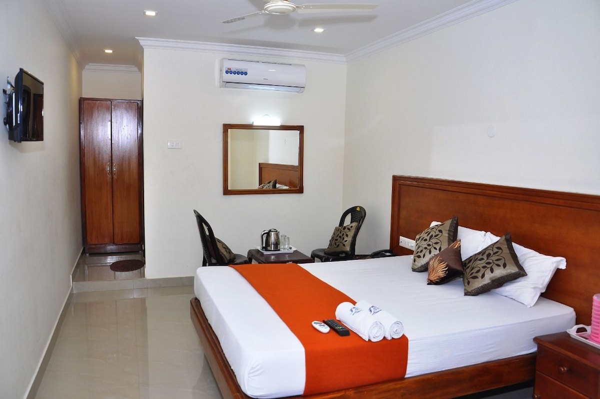 A well-lit room features a spacious bed with decorative pillows, flanked by two comfortable chairs. A television is mounted on the wall, and a mirrored wall reflects the interior. An air conditioning unit is mounted above the bed, enhancing the room's comfort.