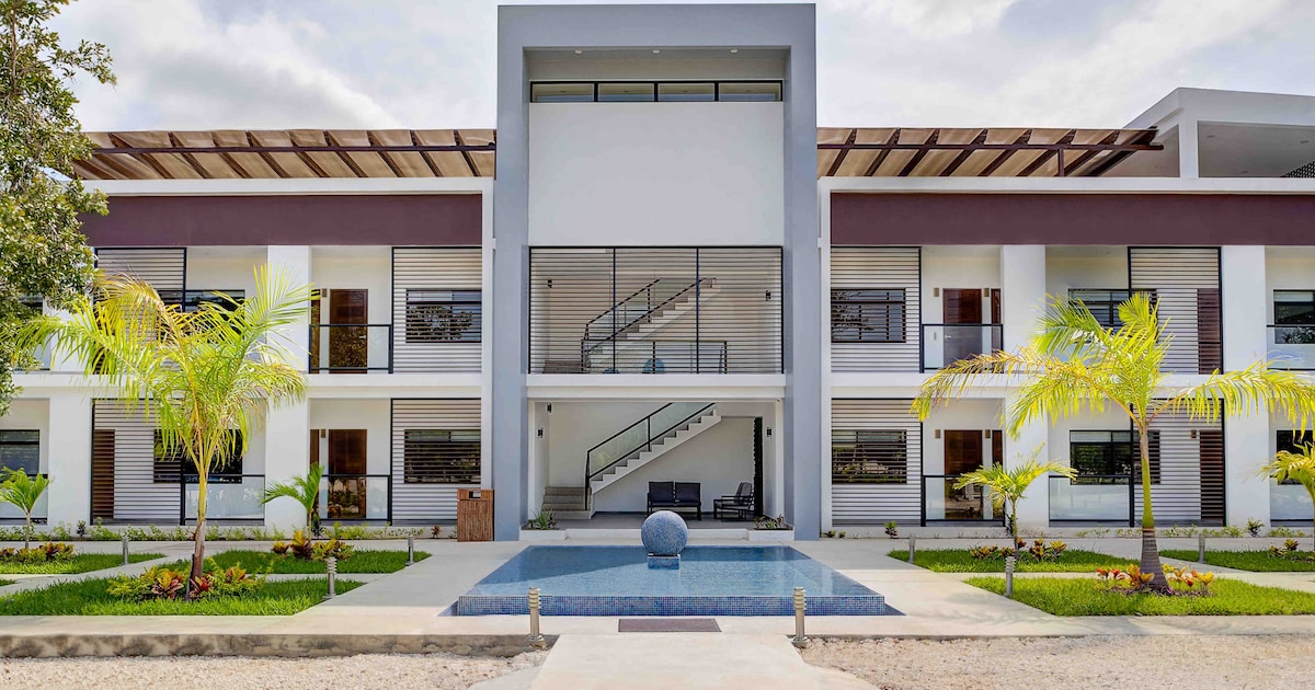 The exterior of a modern two-story boutique hotel is displayed, featuring a large entrance with a minimalist design. Palm trees are positioned in the landscaped garden, complemented by colorful plants. A small water feature reflects the sunlight, enhancing the welcoming space.