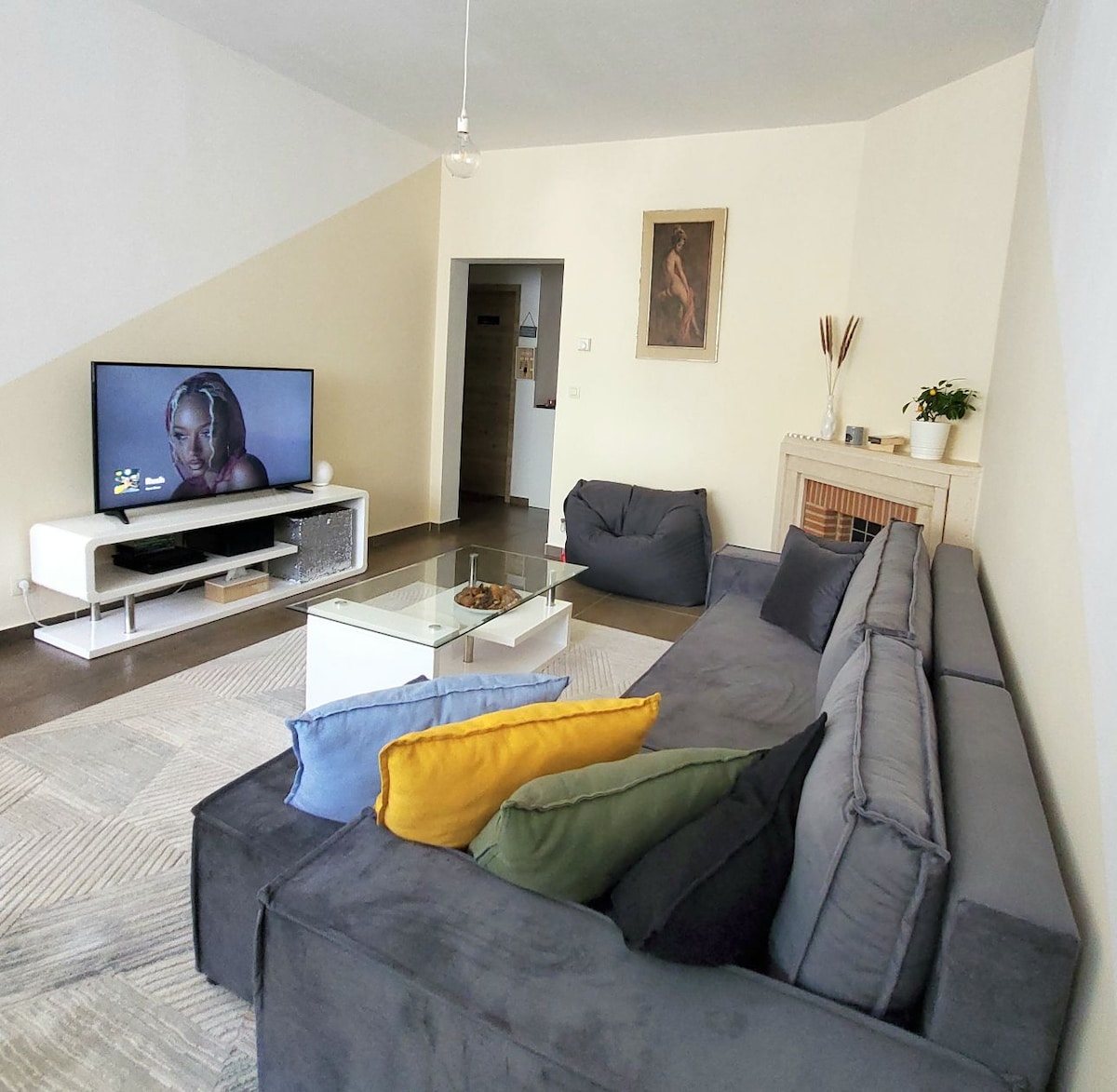A comfortable living area features a large, L-shaped sofa adorned with varied colored pillows. In front, a glass coffee table rests on a textured rug. A television is positioned on a white media console, and artwork adds a touch of decor to the warm-toned walls.