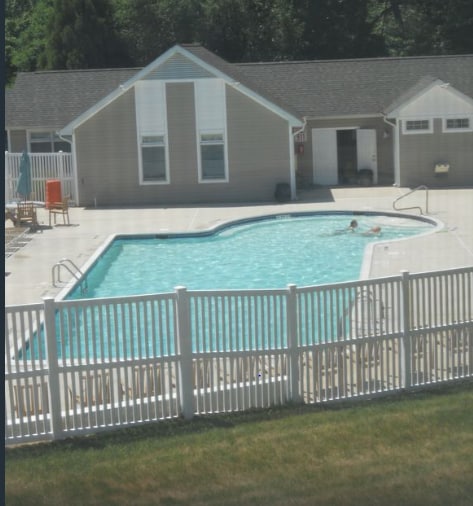 A gated pool area is visible, featuring a curved shape with light blue water and surrounding lounge chairs. Sunlight reflects off the surface, creating a serene atmosphere. The backdrop includes a light-colored building with large windows, further enhancing the tranquil setting.