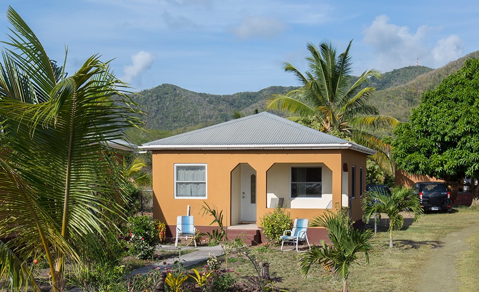 A charming cottage features a light orange exterior and a welcoming front porch. Lush greenery surrounds the property, with palm trees and vibrant landscaping enhancing the tranquil setting. Rolling hills are visible in the background under a clear blue sky.