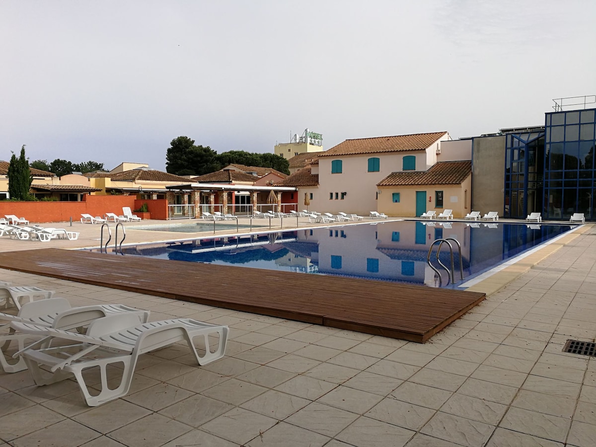 A large outdoor swimming pool is centered within a well-maintained area. Surrounding the pool are several sun loungers arranged on a tiled deck. The area features resort-style buildings in the background, showcasing a blend of modern and traditional architecture.