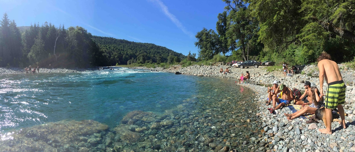 A rocky riverbank is lined with individuals enjoying the clear blue waters of the Rio Azul. Lush greenery and trees rise in the background, and vehicles are parked nearby. Sunlight casts a warm glow on the scene, highlighting the natural setting.