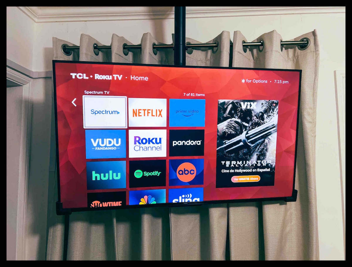 A wall-mounted flat-screen Smart TV displays various streaming options, including popular services like Netflix and Hulu. The screen is positioned above a set of light-colored curtains, creating a functional entertainment space within the room.