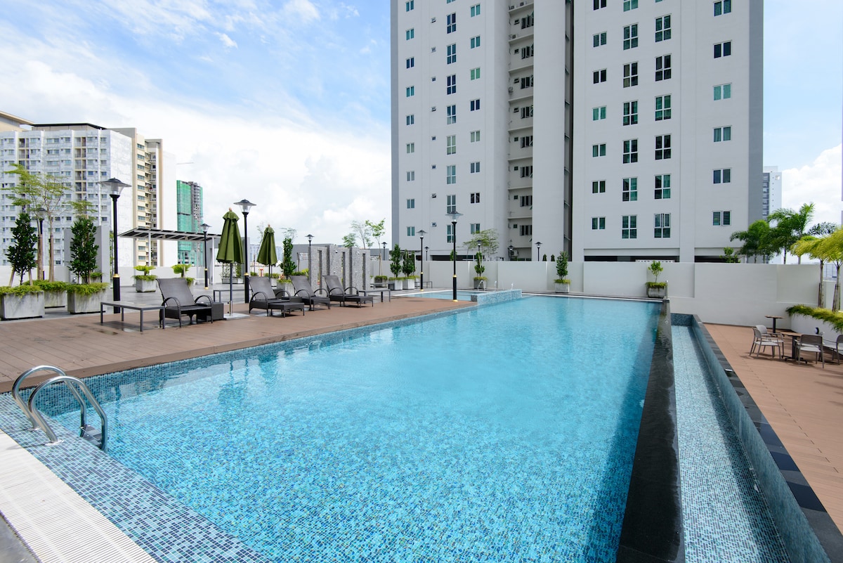 An infinity pool is set within a modern outdoor space, surrounded by sun loungers and umbrellas. The building's tall structure is visible in the background, and a clear blue sky enhances the serene atmosphere.