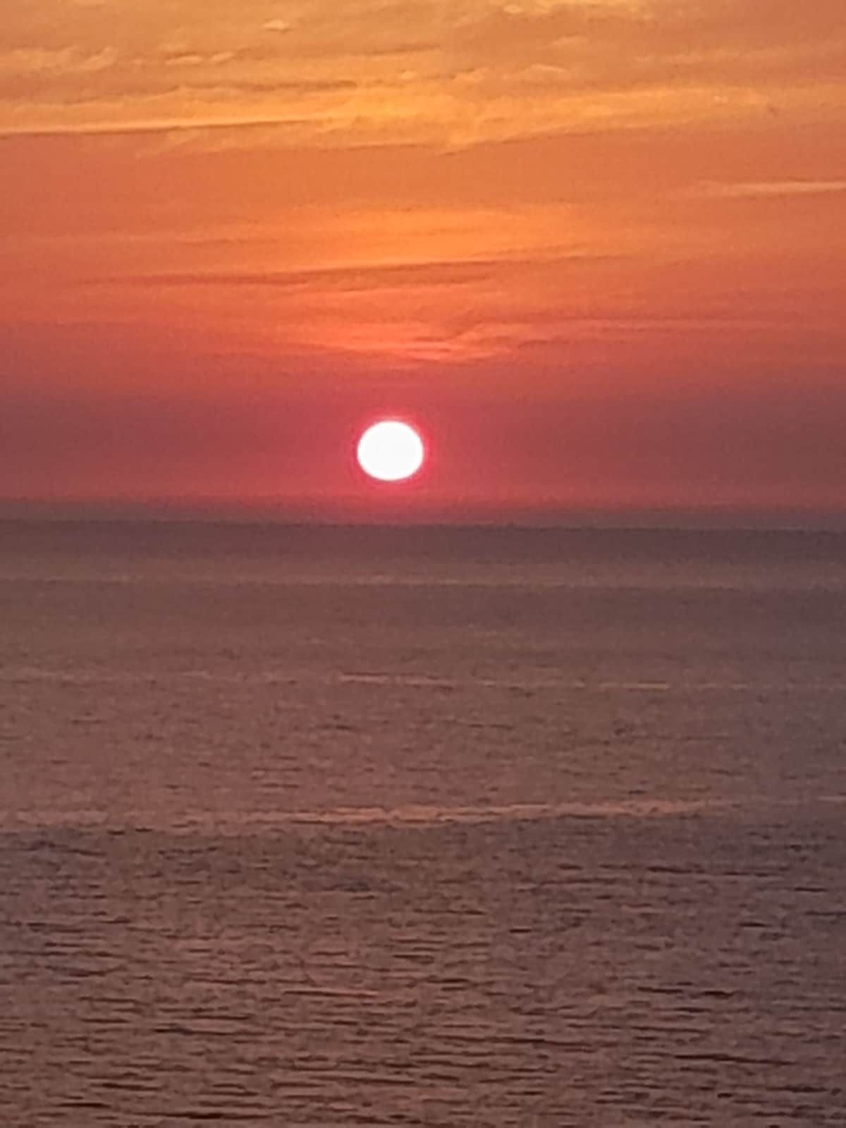 A vibrant sunset is captured over the calm sea, with the sun appearing large and round on the horizon. The sky is filled with warm shades of orange and pink, creating a serene and picturesque view.