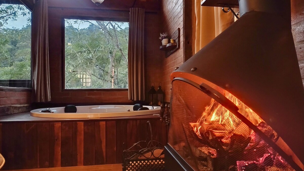 A warm, inviting interior features a spacious hot tub beside a large window, offering views of the surrounding nature. A cozy fireplace is prominently displayed, with the fire softly illuminating the room, creating a comfortable setting for relaxation.