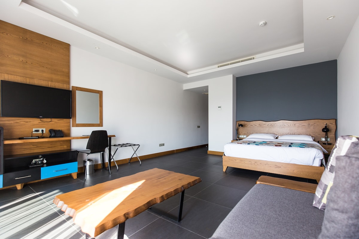 A modern room is furnished with a spacious bed featuring a wooden headboard. A dark accent wall contrasts with the light flooring. A wooden coffee table is positioned in front of a seating area. A flat-screen TV is mounted on the wall, and a desk with a chair is also present.