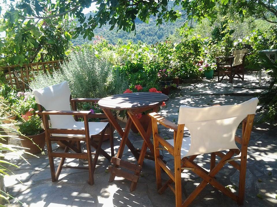 A charming outdoor seating area is surrounded by lush greenery and colorful flowers. Two wooden chairs with white fabric seating are positioned around a small round table, creating an inviting space amidst the natural beauty of the courtyard.