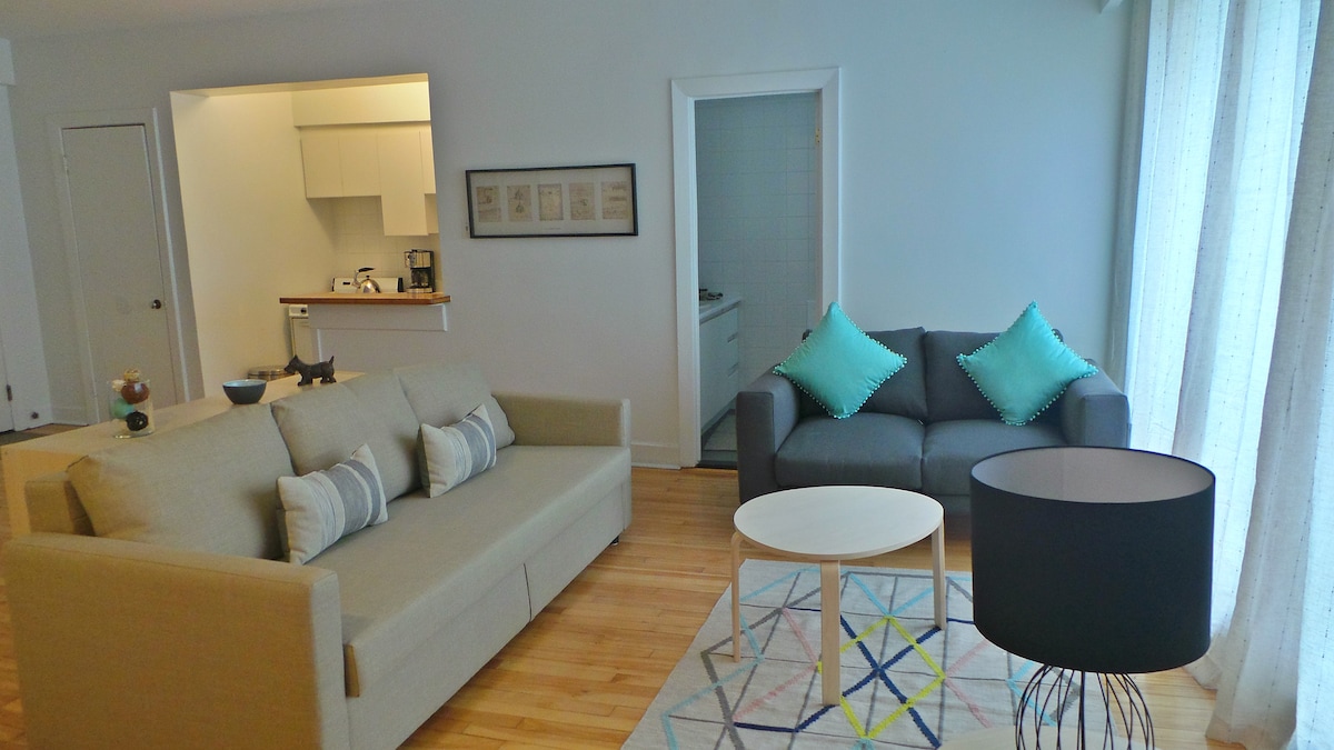 A spacious living area is showcased, featuring a light-colored sofa and a dark gray couch, both adorned with turquoise cushions. A round table sits in the center, accompanied by a floor lamp with a black shade. Natural light enters through sheer curtains, enhancing the room's ambiance.