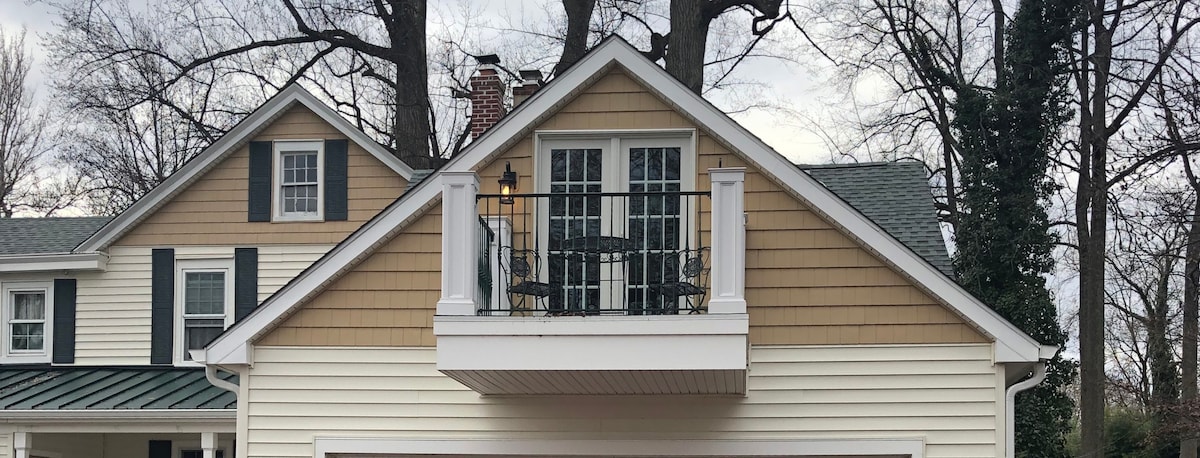 A charming balcony is visible from the loft, featuring two chairs and a small table. The balcony is enclosed by a decorative railing, and framed by tall trees in the background, offering a sense of privacy and connection to nature.