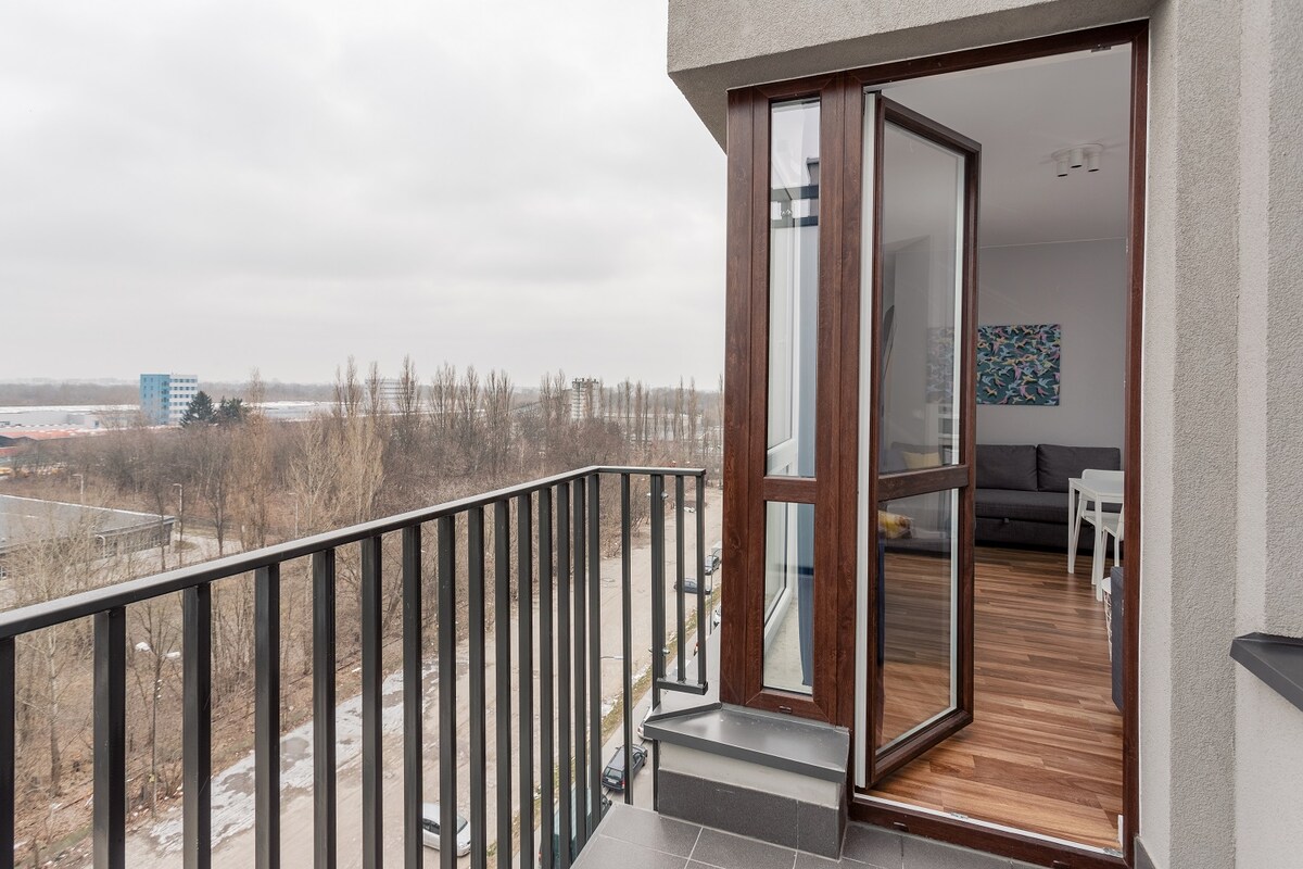 A balcony is visible, featuring a wooden railing and a door leading into an indoor space. The view includes an open area with trees and distant buildings. The floor is tiled, and natural light can be seen entering from the glass door.