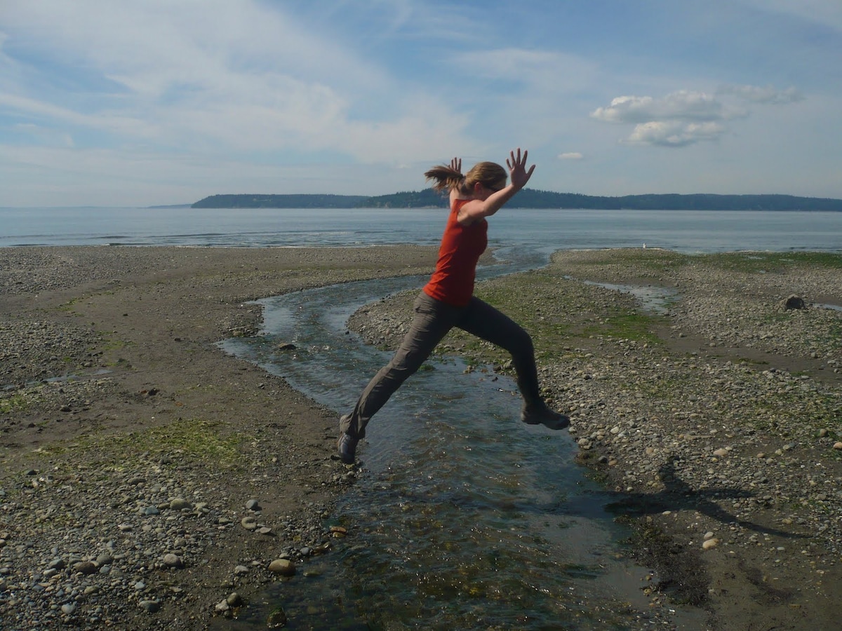 A person is captured mid-air, jumping across a shallow stream that meanders through a rocky beach. The scene features a calm ocean in the background with distant hills under a clear blue sky, suggesting a serene coastal environment.