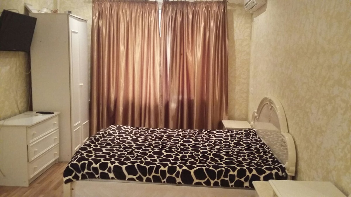 A cozy bedroom features a spacious bed, covered with a patterned black and white blanket. Golden curtains frame a window, allowing natural light to illuminate the room. A white dresser and storage cabinet are positioned against the wall, offering ample space for belongings.