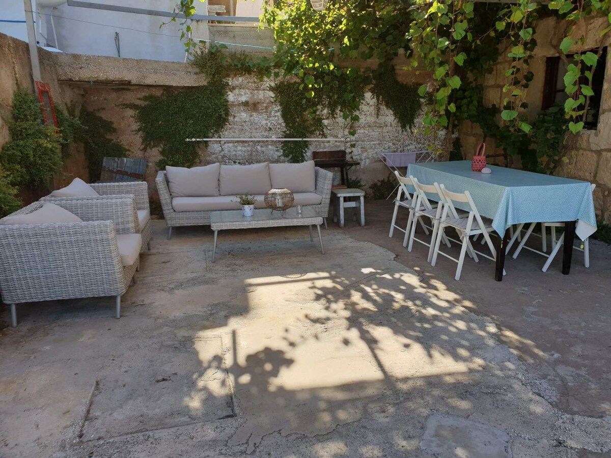 A cozy outdoor terrace is surrounded by stone walls, featuring a relaxed seating arrangement with a sofa and armchairs alongside a dining table. Soft shadow patterns are cast on the ground from the overhead vines, creating a tranquil atmosphere for outdoor meals.