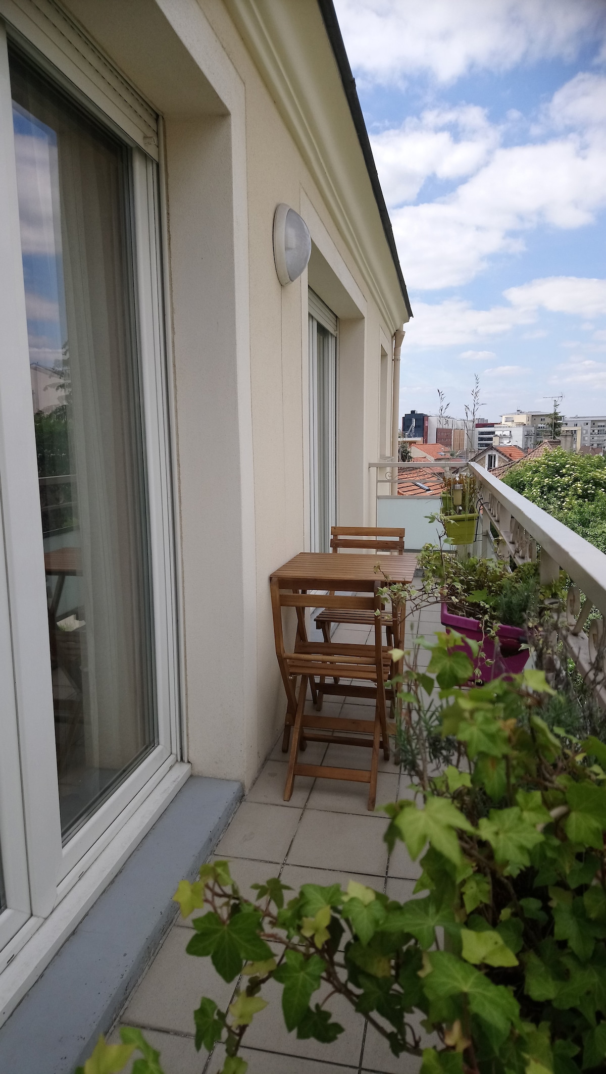 A private balcony is presented, featuring a small wooden table and two folding chairs. Lush greenery is seen in pots along the railing. Large glass doors invite natural light, and the sky above is partly cloudy.