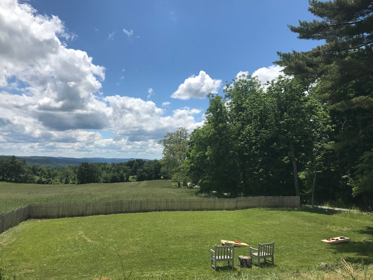 An expansive green yard is enclosed by a wooden fence, featuring two wooden benches positioned for enjoying views of the rolling hills. Large trees provide shade, and a clear blue sky is dotted with white clouds.
