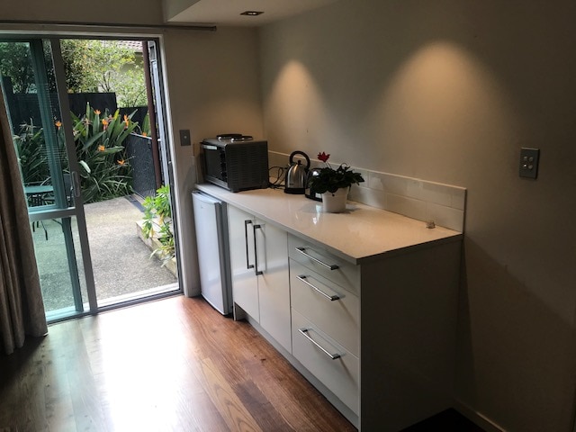 A kitchenette is presented with modern cabinetry and a countertop, featuring an electric kettle and a small oven. A refrigerator is positioned beside the cabinetry, and natural light enters through a ranch slider, illuminating the space and offering a view of a private courtyard with greenery.