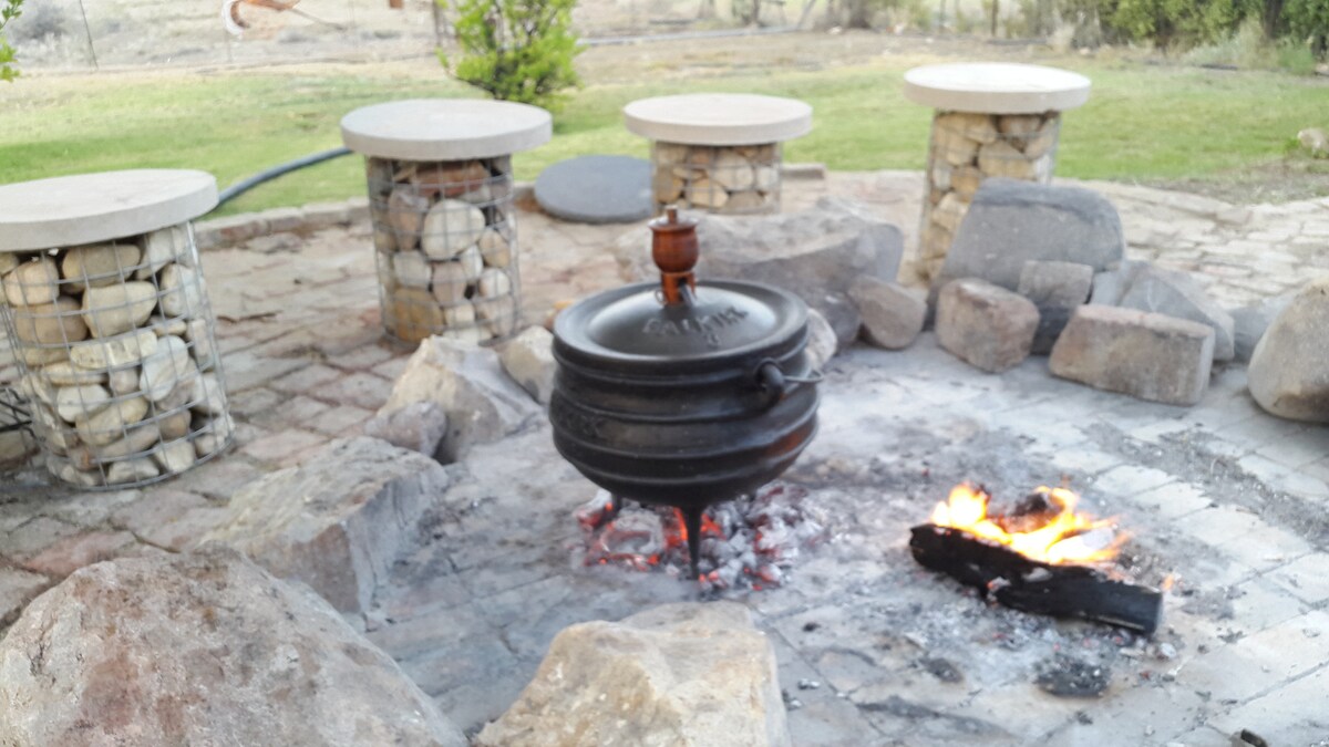 An outdoor fire pit area is showcased with a traditional pot placed over the flames. Surrounding the fire pit are several stone and circular seating arrangements, creating a cozy atmosphere for gatherings. Natural stones and grassy areas enhance the rustic charm.