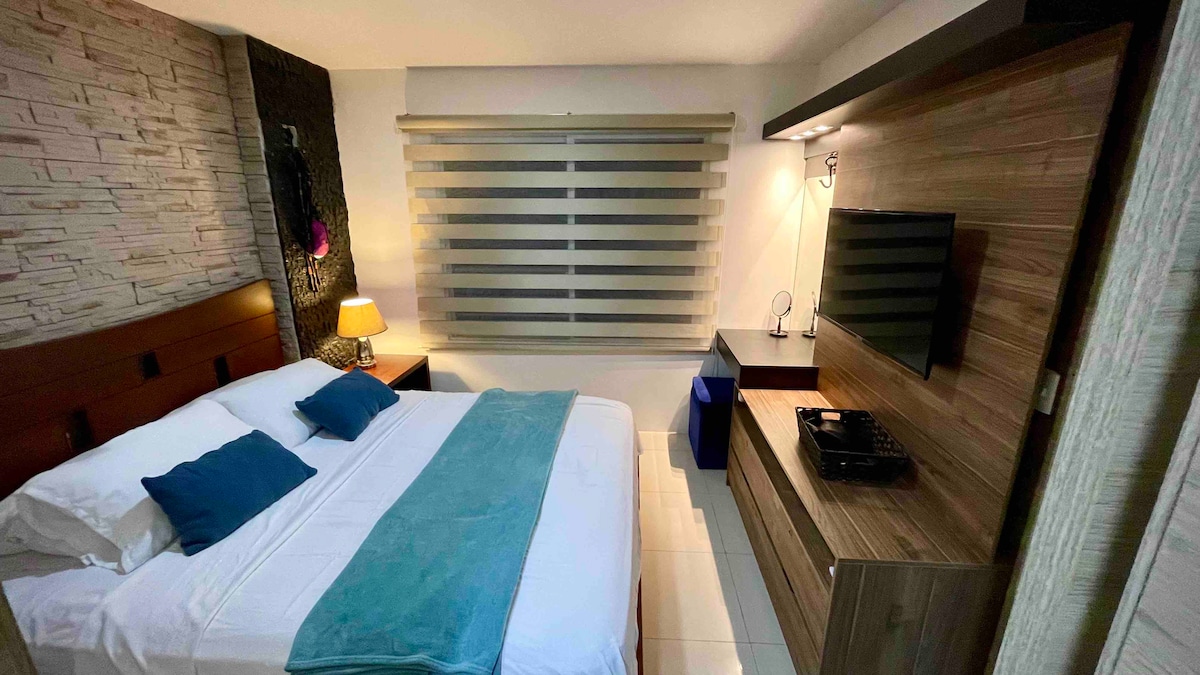 A modern bedroom features a spacious bed with a light blue throw and decorative pillows. A flat-screen TV is mounted on a wooden panel, and natural light filters through the contemporary striped window shades. A bedside lamp adds subtle illumination to the inviting space.