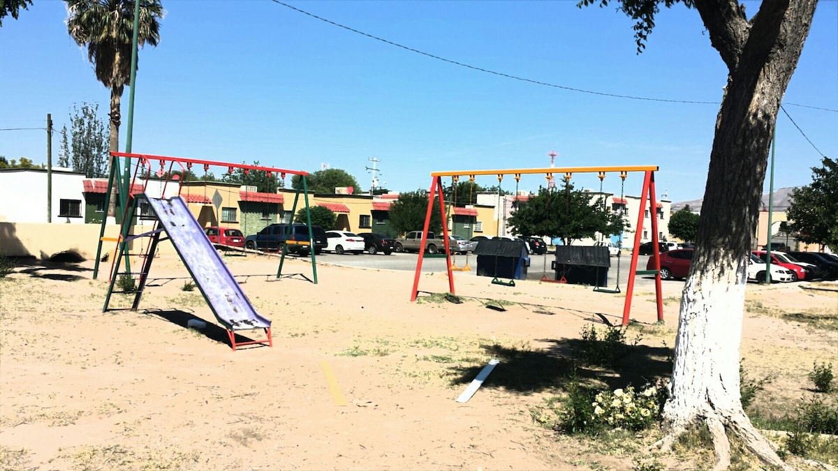 An outdoor playground is seen featuring a slide and two swing sets. The area is surrounded by sand and grass, with trees providing some shade. Nearby buildings can be observed in the background, complementing the recreational space.