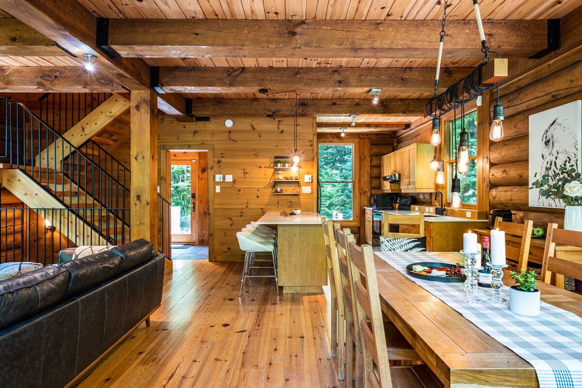 A spacious, wooden interior features exposed beams and natural light flooding the area. A long dining table is set for meals, complemented by a cozy couch nearby. The kitchen is visible in the background, offering an inviting space for gatherings. The entrance leads outside to greenery.