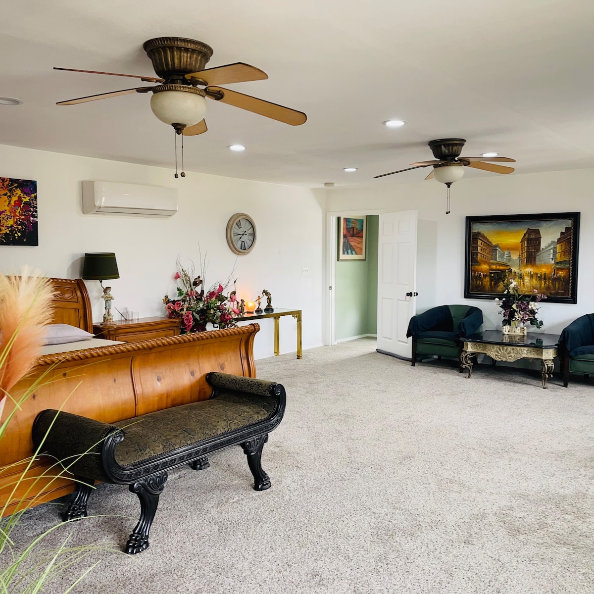 A spacious living area features two ceiling fans, a comfortable king bed with decorative furnishings, and a stylish bench at the foot. Lively floral arrangements and wall art add character, while soft carpet covers the floor, enhancing the room's cozy ambiance.