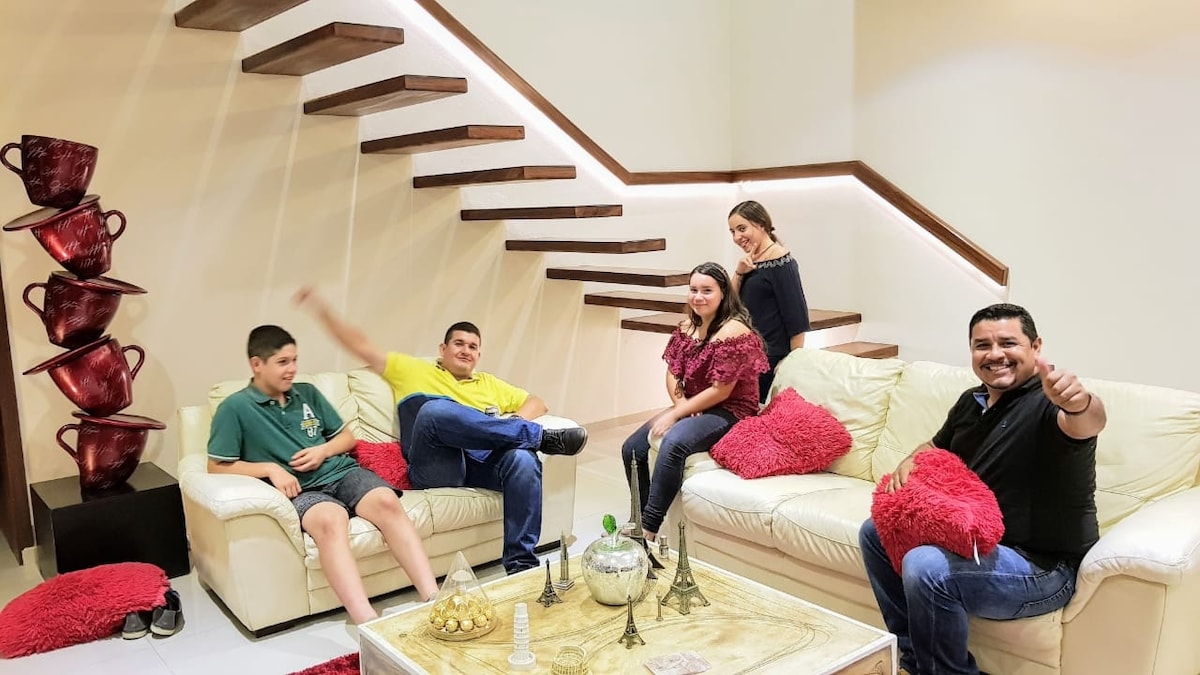 A spacious living area features a light-colored sofa adorned with red cushions. A glass coffee table sits at the center, with decorative items placed on it. A staircase with wooden steps is visible in the background, complemented by soft lighting.