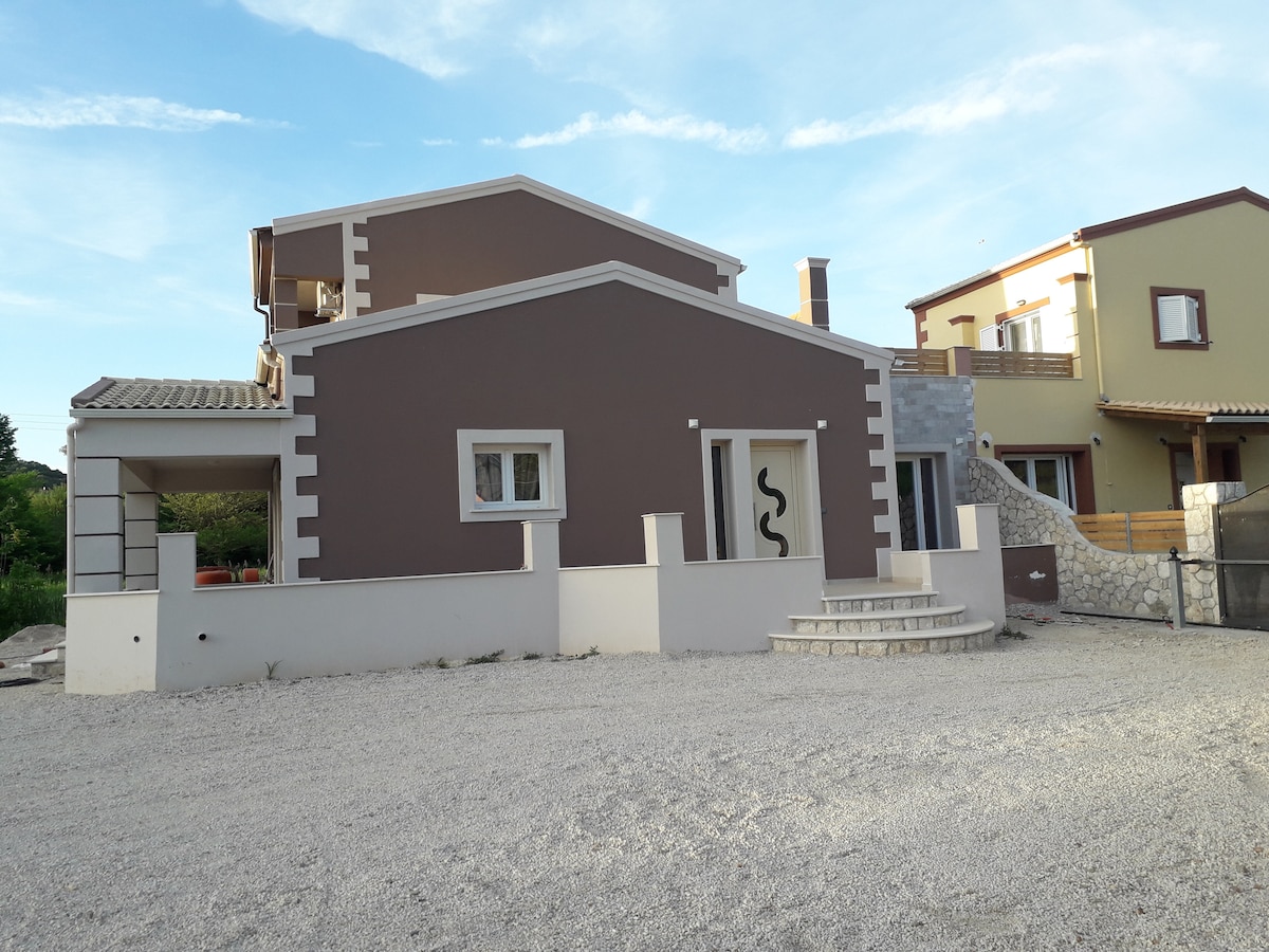 The villa features a modern design with a combination of brown and stone exteriors. A set of stone steps leads to the entrance, framed by a simple decorative element. Surrounding gravel provides a spacious setting, complemented by lush greenery in the background.