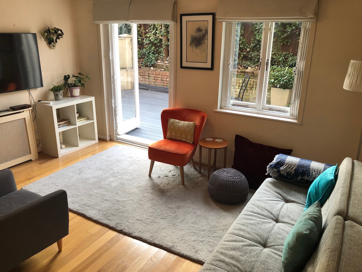 A living room area features a grey sofa and a comfortable orange armchair, positioned for relaxation. A small side table and a pouf are nearby. Natural light enters through double doors leading to a private outdoor space, with greenery visible beyond.