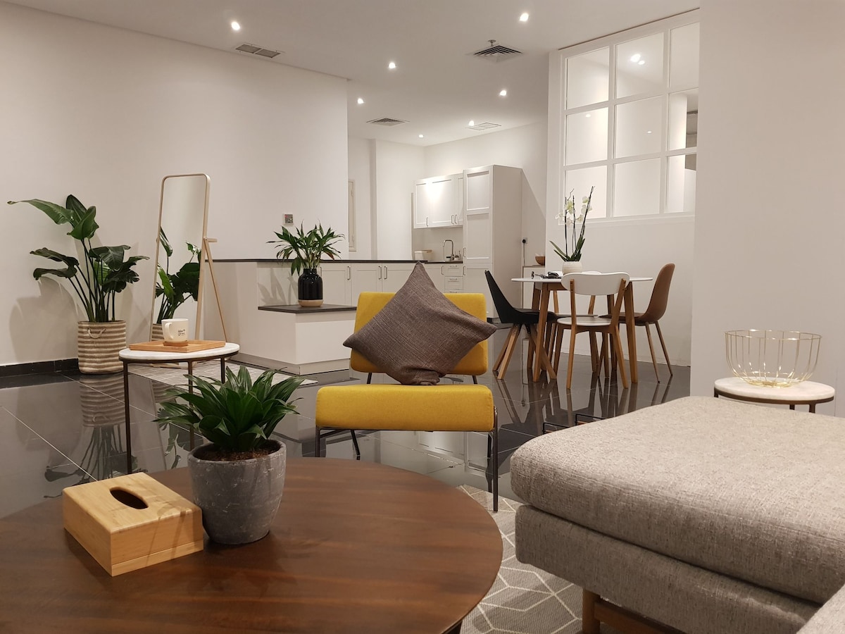 A modern living space is displayed, featuring sleek furniture, including a gray sofa and a yellow accent chair. Indoor plants are strategically placed, adding greenery to the atmosphere. A dining table with wooden chairs and a well-equipped kitchen area are visible in the background.