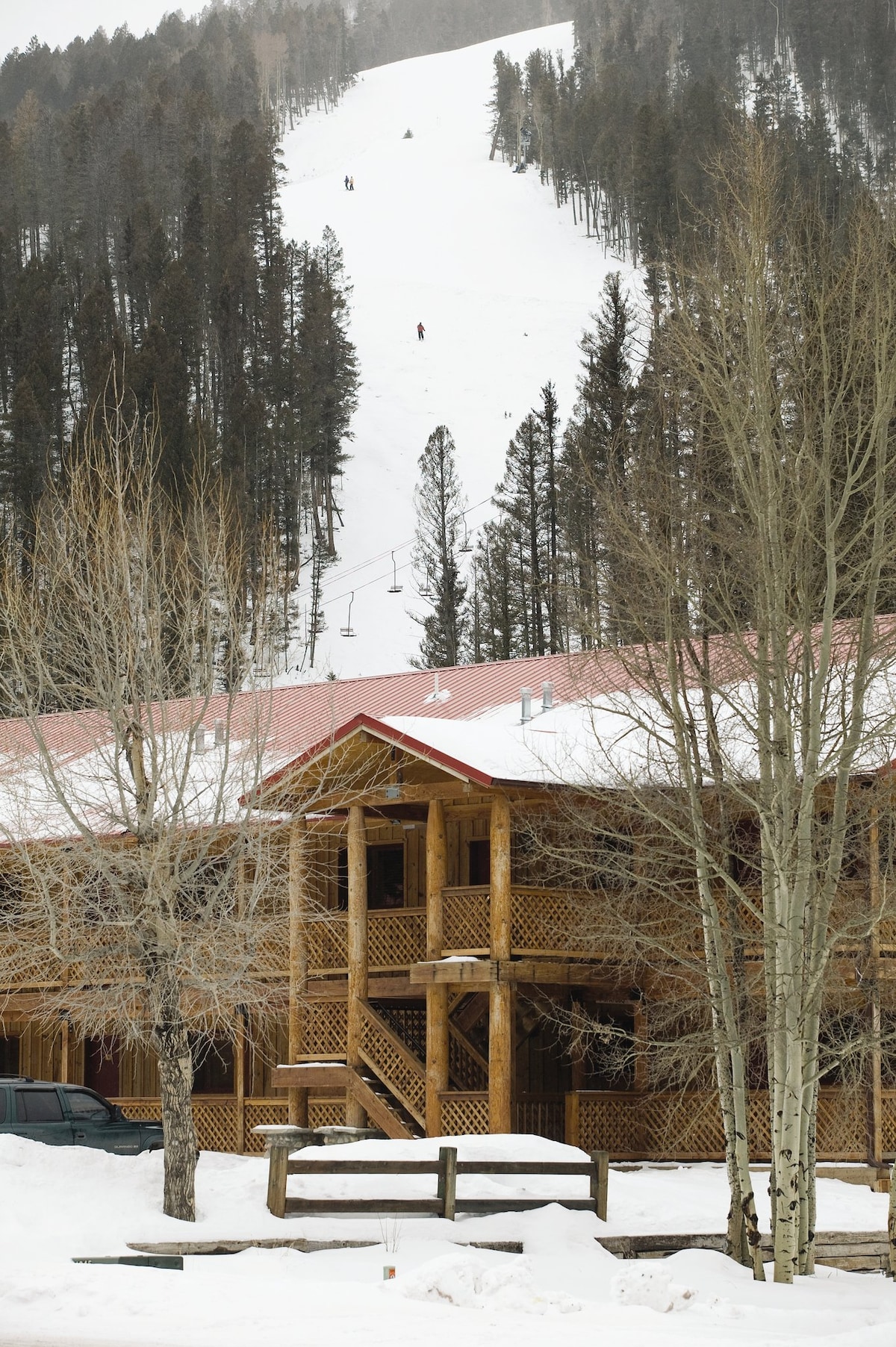 A lodge with a wooden exterior is framed by leafless trees and snow-covered ground. The ski slope is visible in the background, with skiers navigating down the hill. The building features multiple levels and a red roof, complementing the winter landscape.