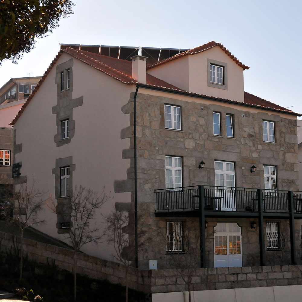 The exterior of a multi-story building is displayed, featuring a mix of stone and plaster. Balconies with black railings are visible on the lower level, while a red roof adds contrast. Windows are aligned symmetrically, complementing the overall architecture.