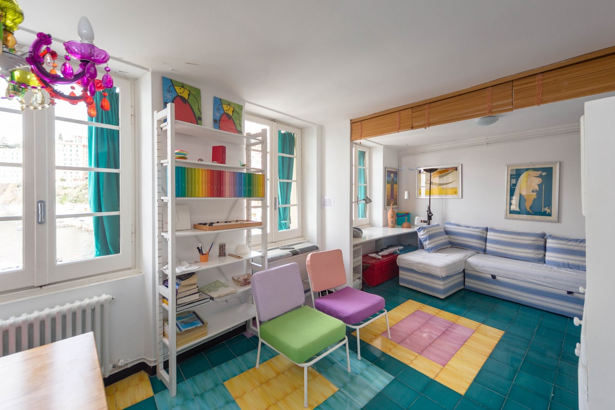 A welcoming living area features a cozy seating arrangement with a spacious sofa and colorful chairs in pink and green. A bright bookshelf holds various items, while windows with turquoise curtains allow natural light to fill the room. The vibrant tiled floor adds a cheerful touch.