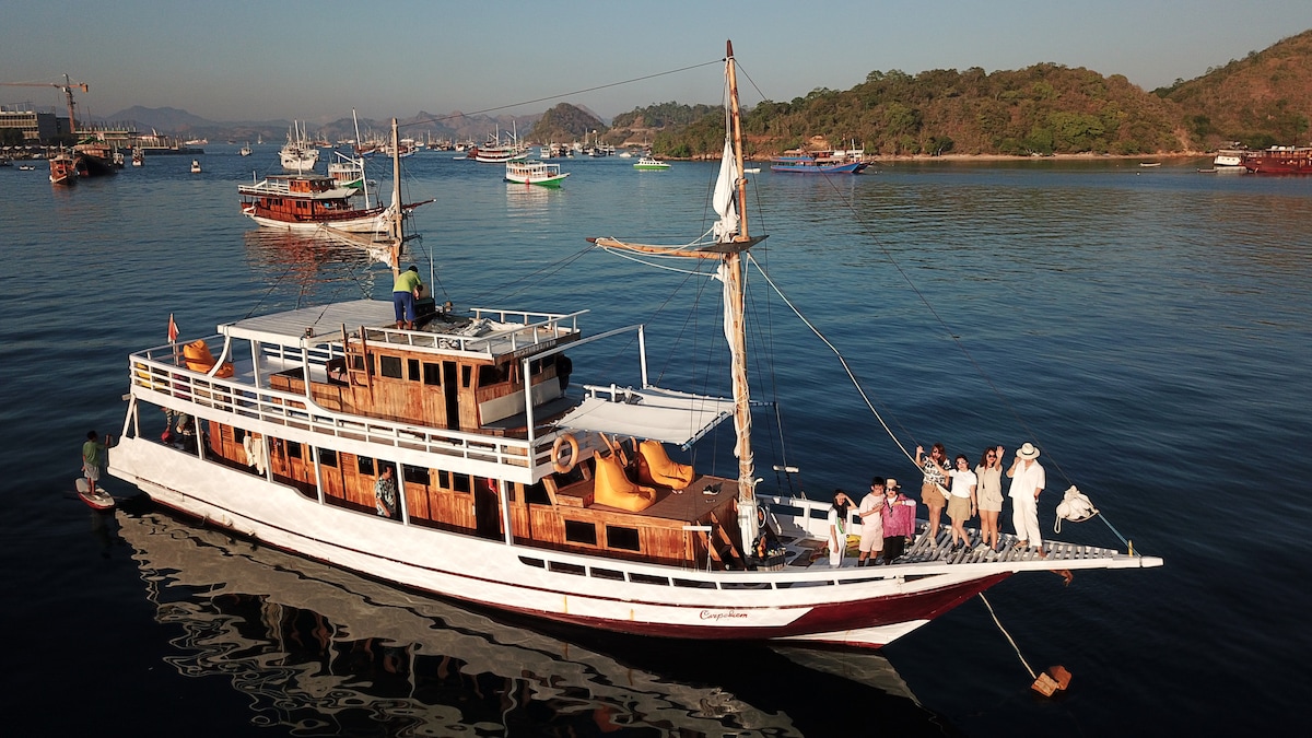 The boat, Carpediem, features a traditional wooden design with a spacious upper deck for sunbathing. Guests are seen enjoying their time on board, surrounded by calm waters and distant hills, creating a serene atmosphere conducive to relaxation and exploration.