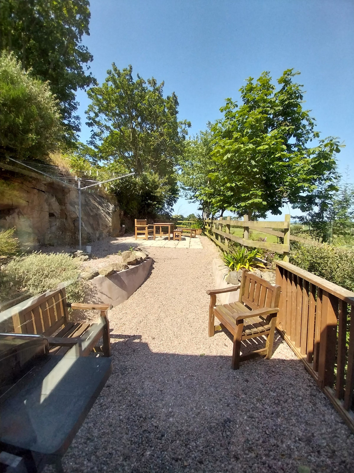 A gravel pathway lined with greenery leads towards a secluded outdoor area. Two wooden benches face a sunlit patio space, surrounded by trees and shrubs, creating a tranquil environment. A wooden fence marks the boundary, providing a sense of privacy.