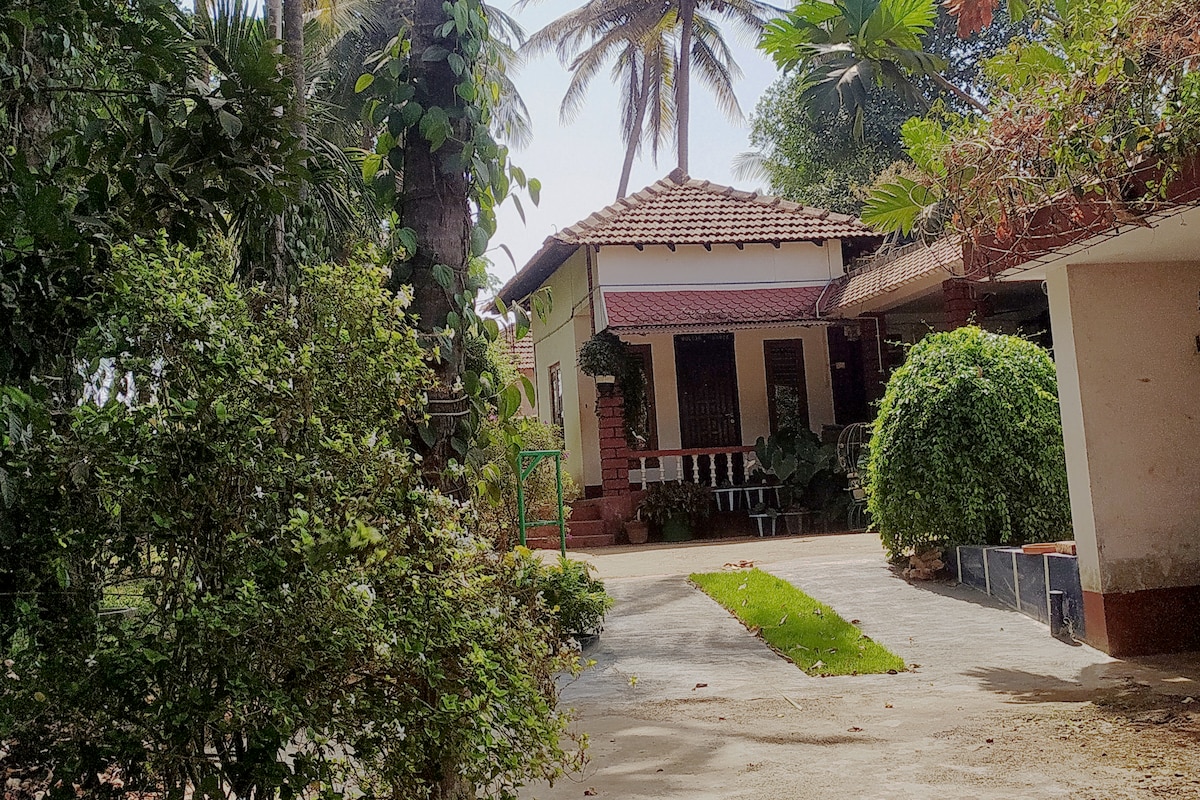 A single-story house is framed by lush greenery, featuring a red-tiled roof and a welcoming front porch. Pathways lead through well-maintained gardens, with tropical plants and trees, creating a serene atmosphere. The landscape offers shade and tranquility, inviting guests to relax outdoors.