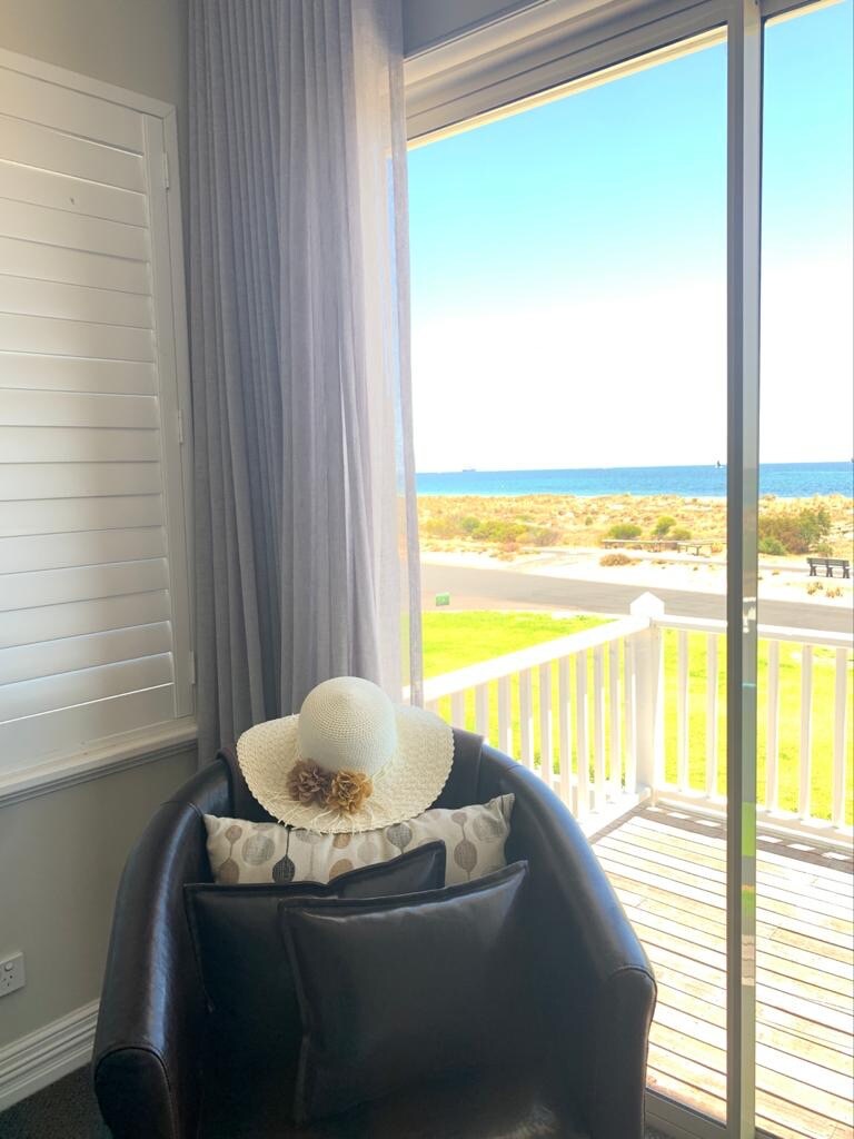A cozy armchair sits beside large glass doors, offering a view of the ocean. A wide-brimmed hat rests on decorative pillows, complementing the light, airy decor. Sheer curtains allow natural light to fill the space, enhancing the inviting atmosphere.