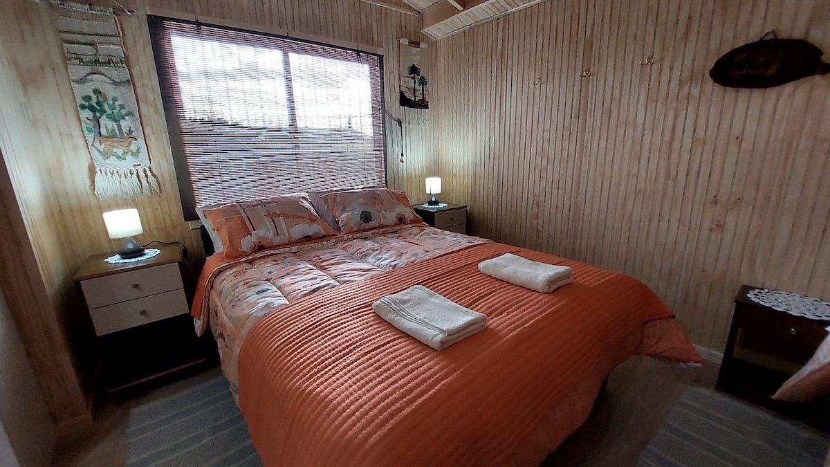 A cozy bedroom features warm wooden walls and a large window allowing natural light. A spacious bed is adorned with orange bedding, complemented by neatly arranged towels. Two bedside tables with lamps are situated on either side, providing subtle illumination.