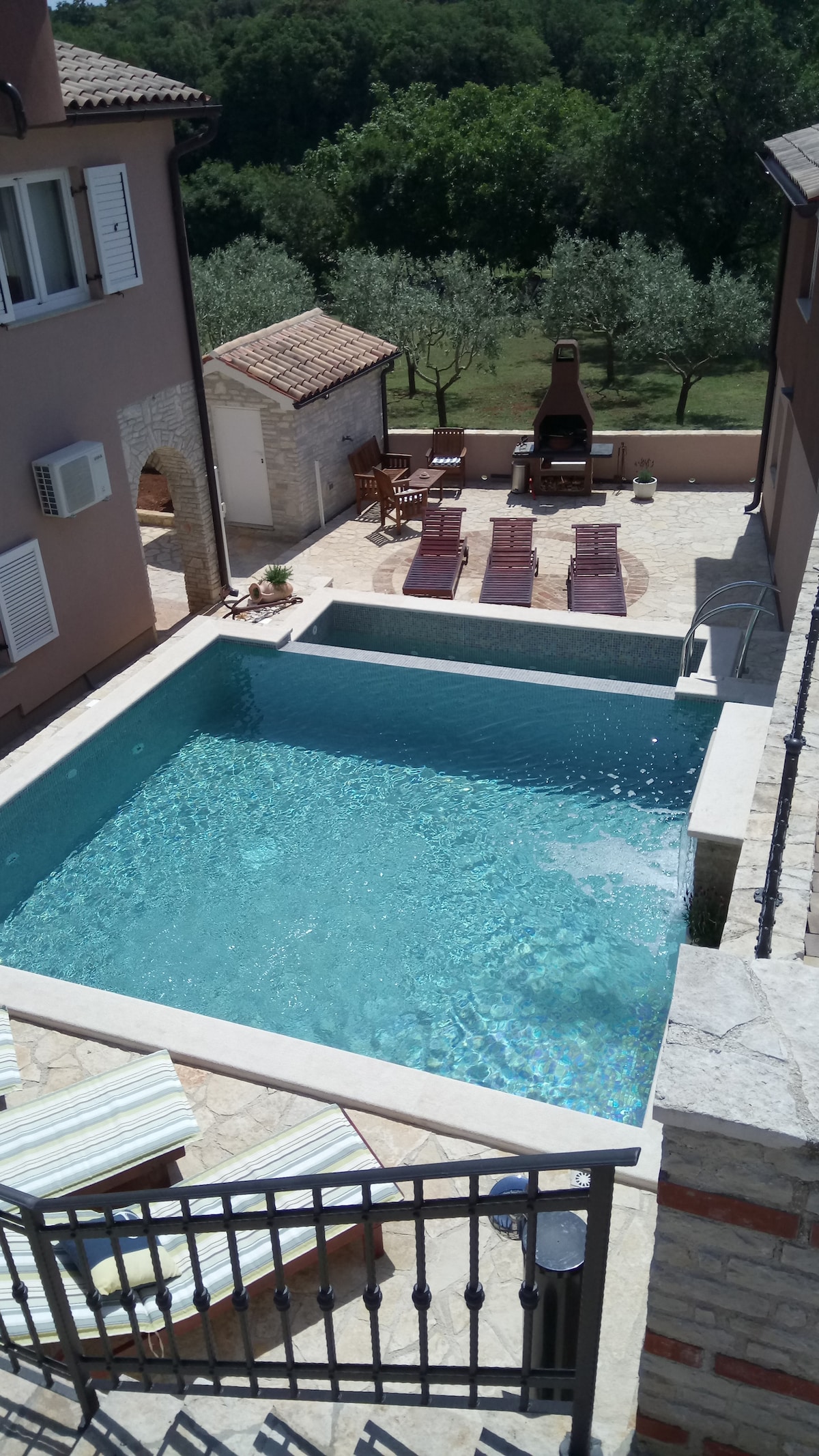 A private swimming pool is surrounded by a spacious stone patio featuring several lounge chairs for relaxation. An outdoor fireplace is positioned nearby, complemented by a serene backdrop of olive trees. The area is well-lit by natural sunlight, enhancing the peaceful setting.