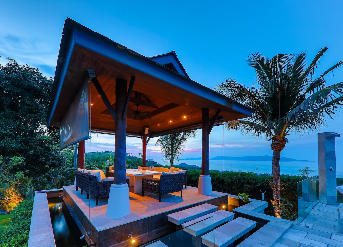 An outdoor sala with a thatched roof features two wicker lounge sofas and a coffee table, positioned for serene sea views. Tropical palms stand nearby, while soft lighting highlights the space during twilight. Steps lead down to a lush landscaped area, adding to the ambiance.