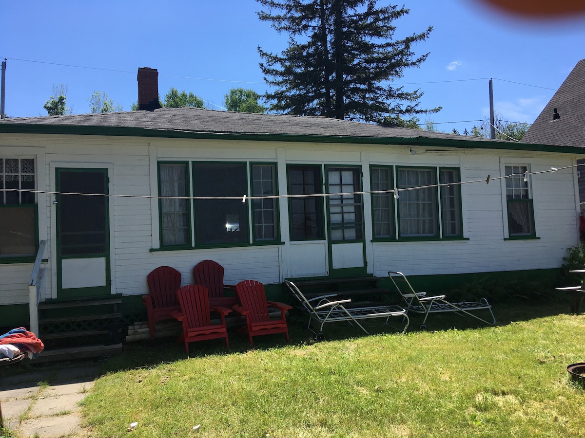The exterior of a cottage is presented, featuring a long porch with multiple windows framed in green. A set of red Adirondack chairs and lounge chairs are placed on a grassy area, with a clothesline visible in the background under a clear blue sky.