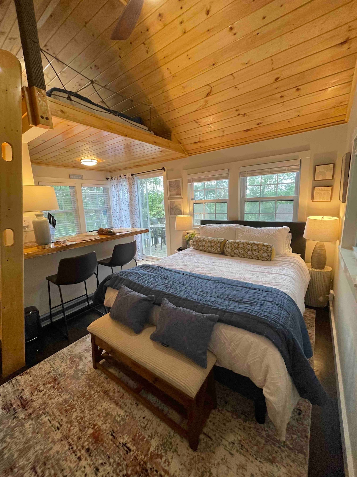 A warm studio cottage features a spacious bed dressed in neutral tones, positioned against a backdrop of wooden paneling. A small kitchen island with bar stools is visible. Large windows allow natural light to fill the room, with soft lighting provided by bedside lamps.