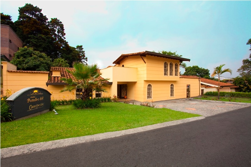 The exterior of Portales Del Campestre is depicted with a warm-colored facade and a tiled roof. Lush green landscaping surrounds the entrance, featuring palm trees and manicured grass. A pebble pathway leads to the door, while an ample driveway provides parking space.