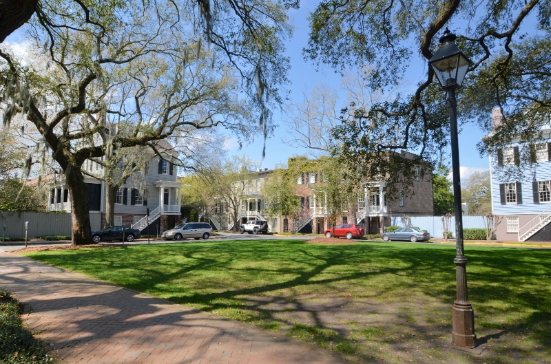 A spacious green square is framed by a variety of historic buildings with classic architectural features. Large oak trees provide shade, while a lamp post stands on the edge of the path, creating a serene atmosphere. Red parked cars are visible along the street.