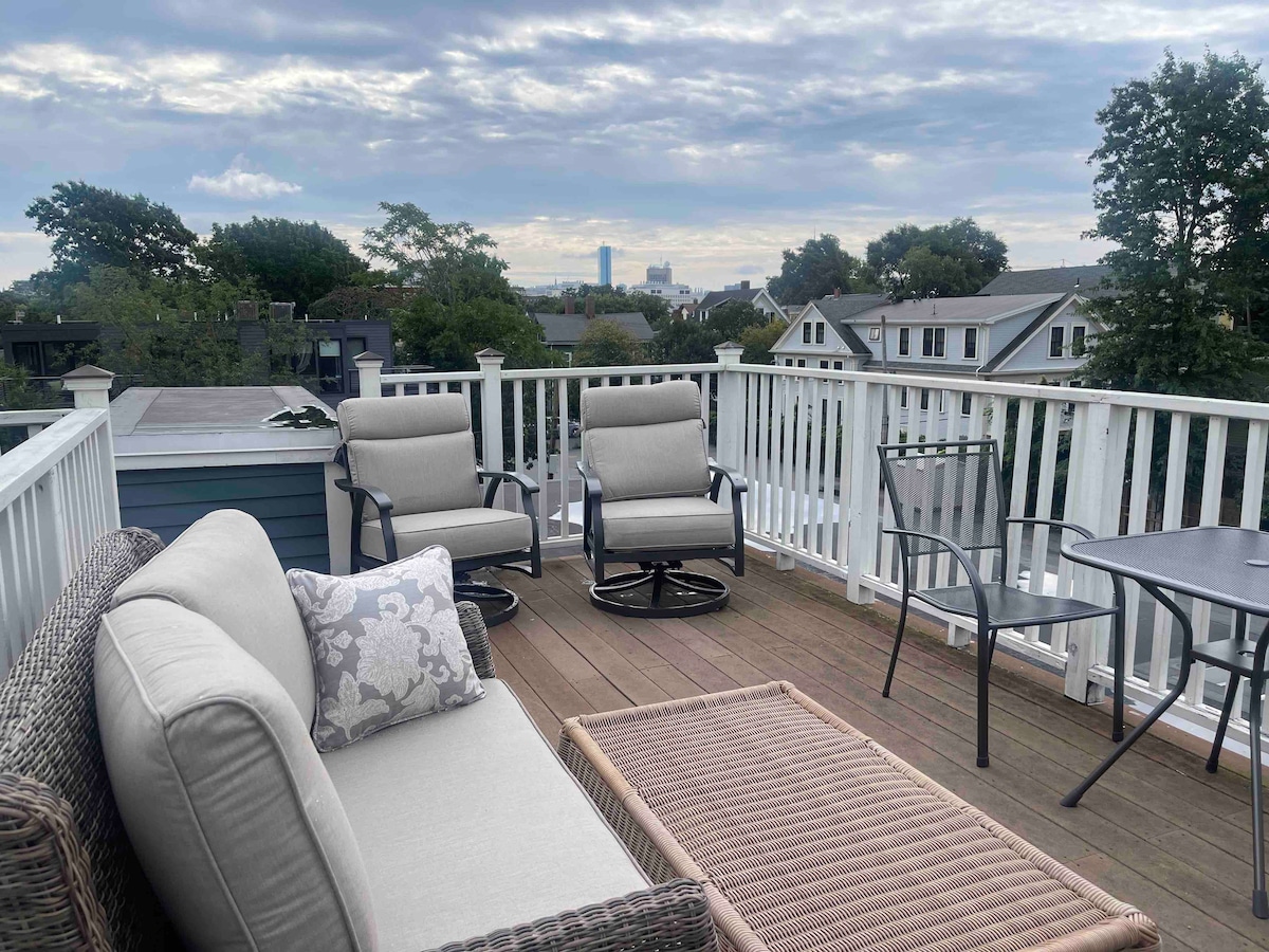 A private rooftop terrace features comfortable outdoor seating arrangements and a table. The space is well-furnished with a sectional sofa adorned with throw pillows, providing a relaxed atmosphere. Views of the surrounding neighborhood and distant city skyline are visible in the background.