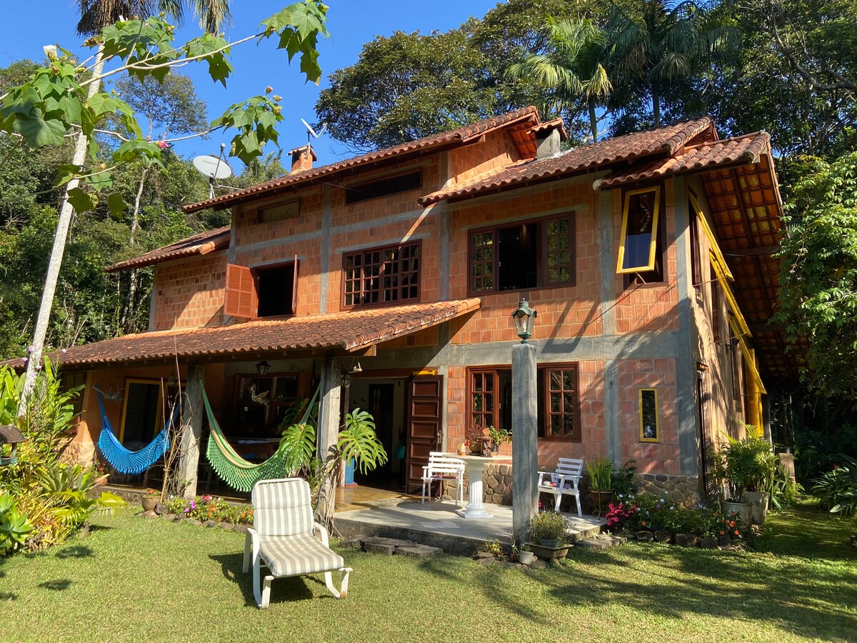 The two-story house is constructed with warm-toned materials, featuring decorative windows and a tiled roof. A spacious entrance area is visible, highlighted by a hammock and outdoor seating. Lush greenery surrounds the property, complementing the serene outdoor setting.