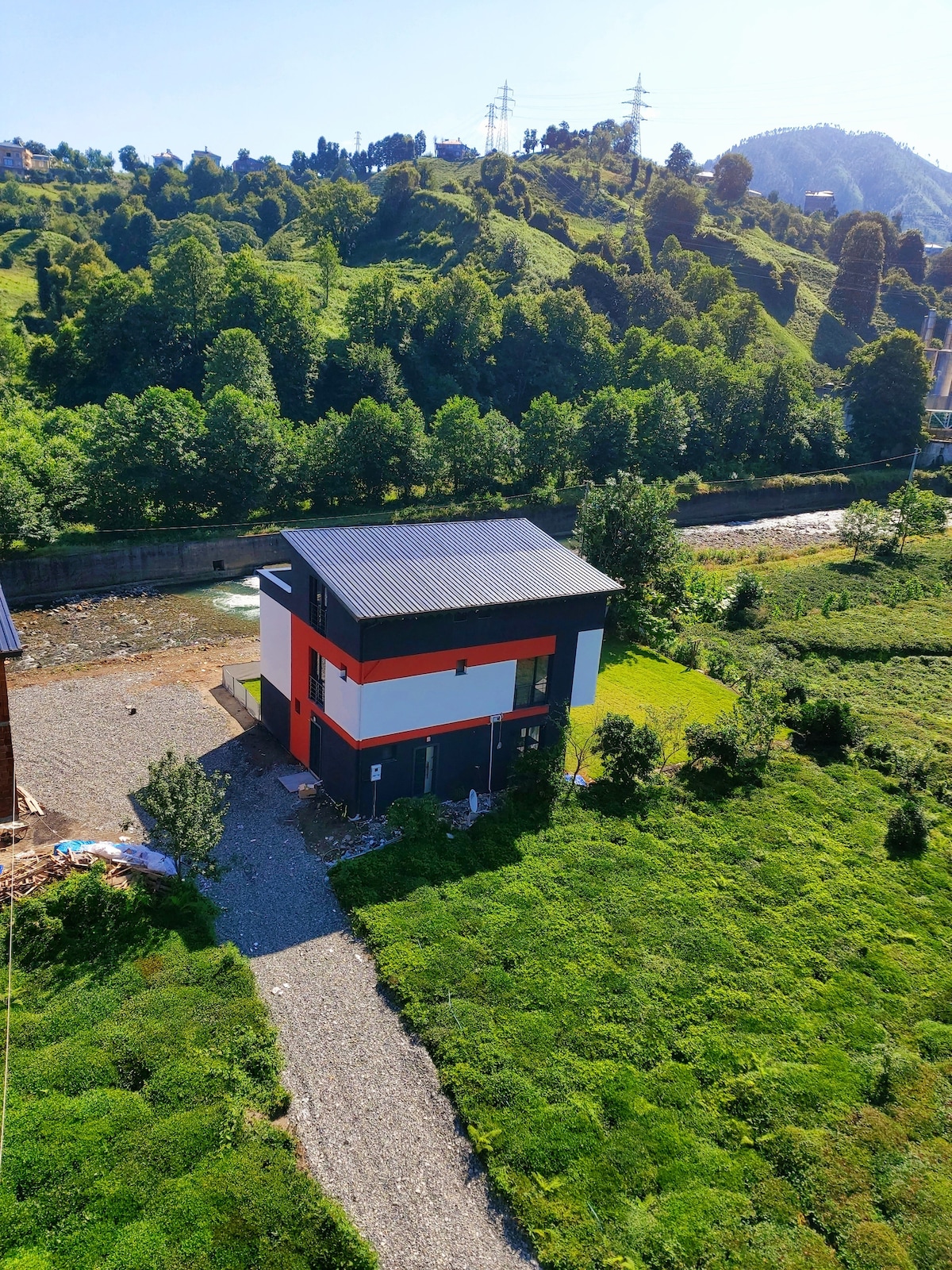 A modern two-story building is surrounded by lush greenery and rolling hills. A pathway leads to the structure, with large windows reflecting the sunlight. A gentle stream flows nearby, enhancing the serene environment.